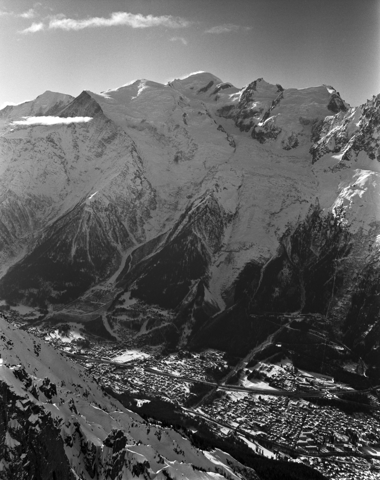 Black and white photograph of Mont Blanc's snow-covered massif towering above a forested valley with a town nestled below.