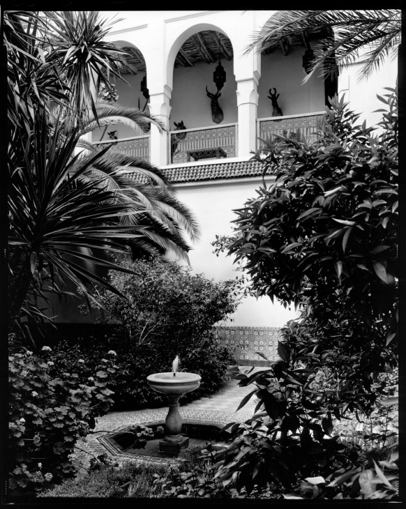 Black and white photograph of a Moroccan riad courtyard with arched doorways, decorative furnishings, and lush tropical plants.
