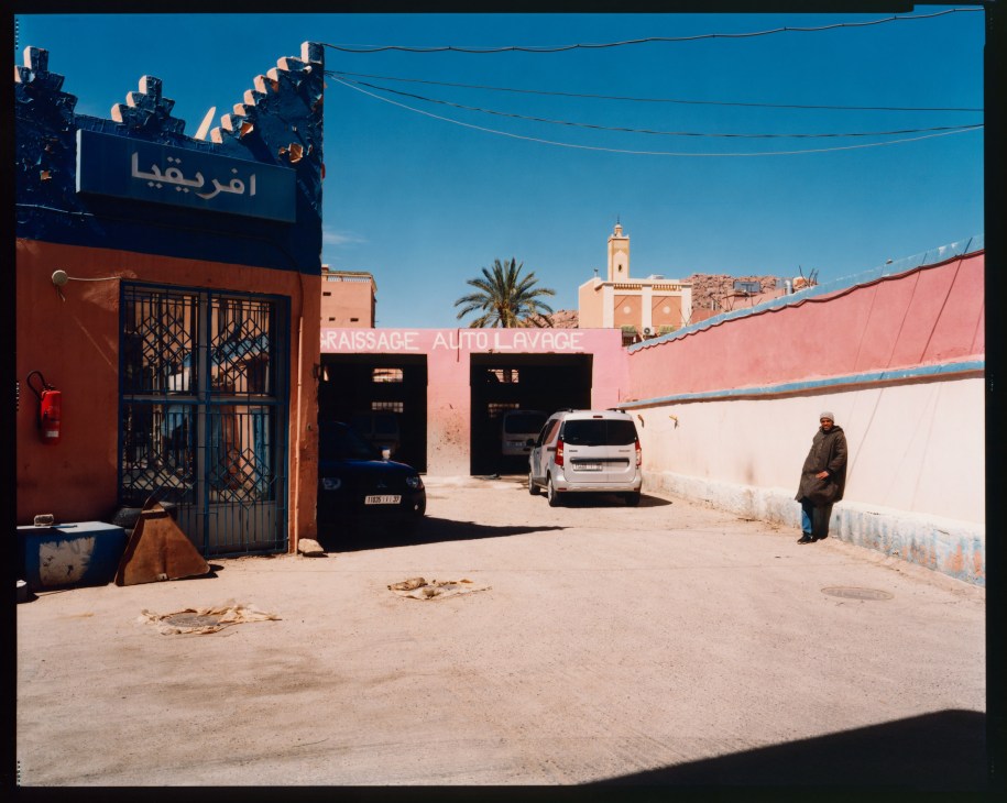 A car wash facility in Tafraoute with colorful red and blue buildings, parked vehicles, and a person standing in the dusty courtyard.