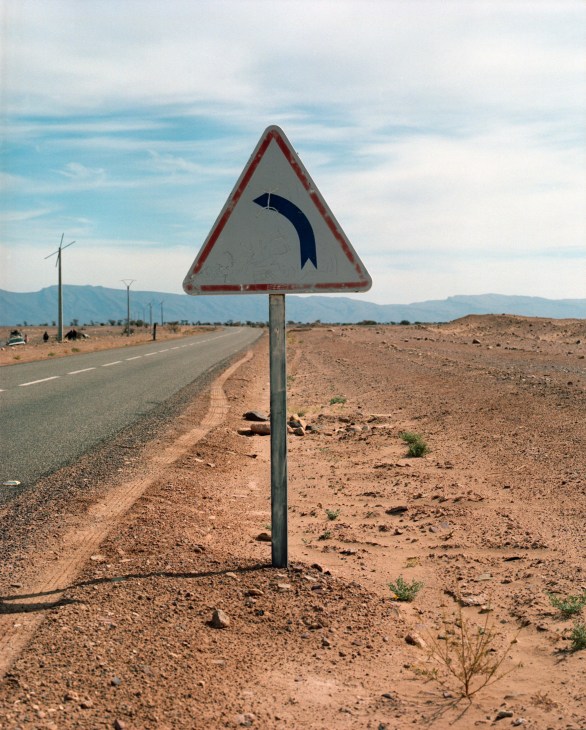 A triangular road sign with a curved arrow symbol on a desert highway in Morocco with mountains in the distance.