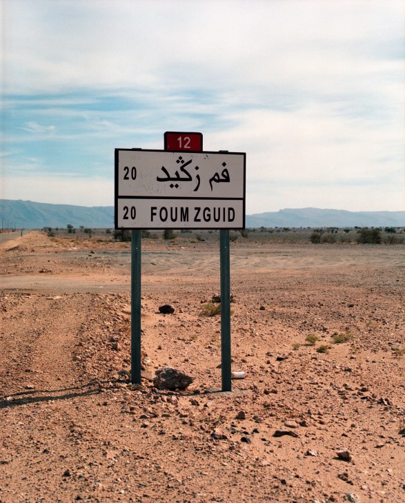 A bilingual road sign for Foum Zguid in Arabic and English standing in an arid desert landscape with mountains in the distance.