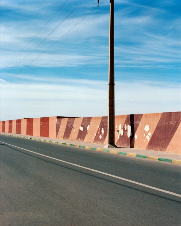 A tall rusty iron pole stands beside a weathered terracotta-colored wall with white paint marks along a Moroccan roadside.