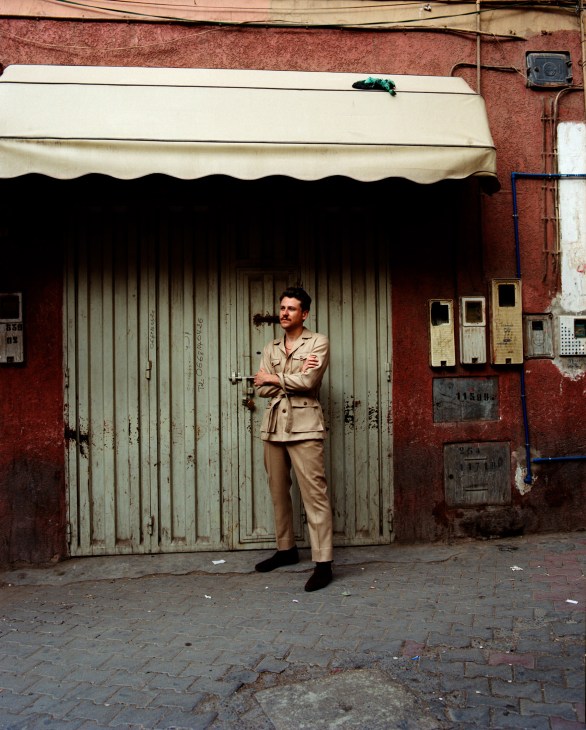 Man in beige suit standing against red wall with corrugated metal door and awning in Morocco.