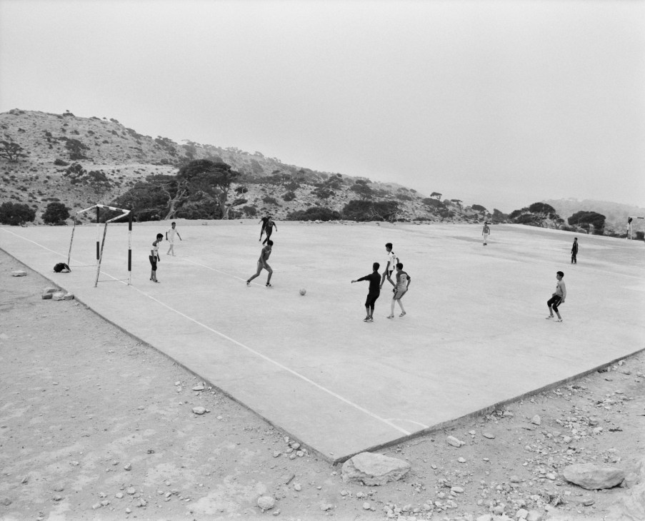 Children playing football on a concrete court with arid hills and scattered trees in the background.