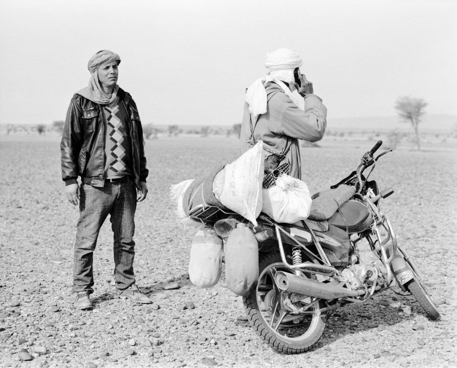 Two men in traditional North African clothing stand with a motorcycle loaded with supplies in a desert landscape.