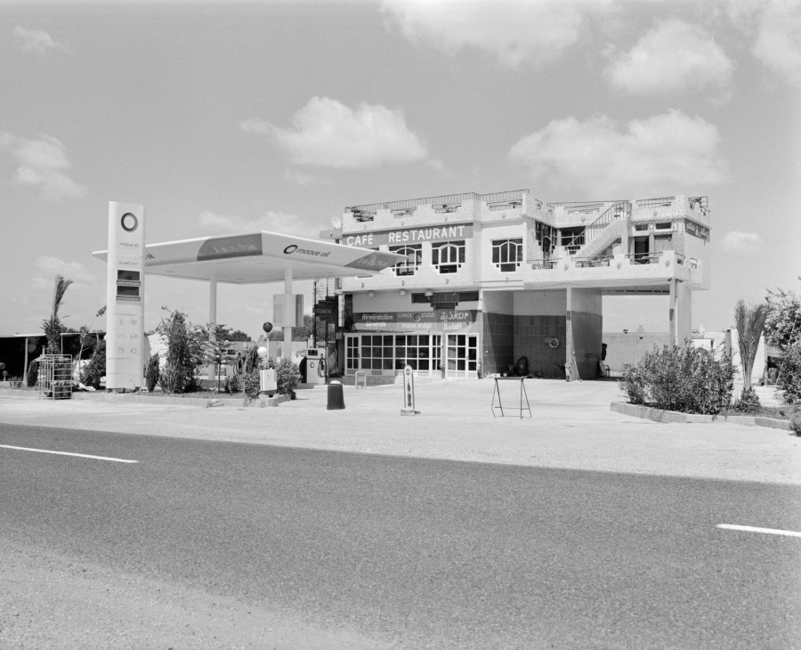 A modernist white gas station and café-restaurant building with geometric lines beside a desert road.