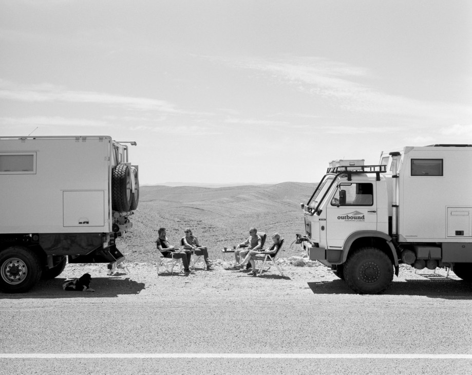Two couples sit in camping chairs between large expedition vehicles on a desert road in Morocco.
