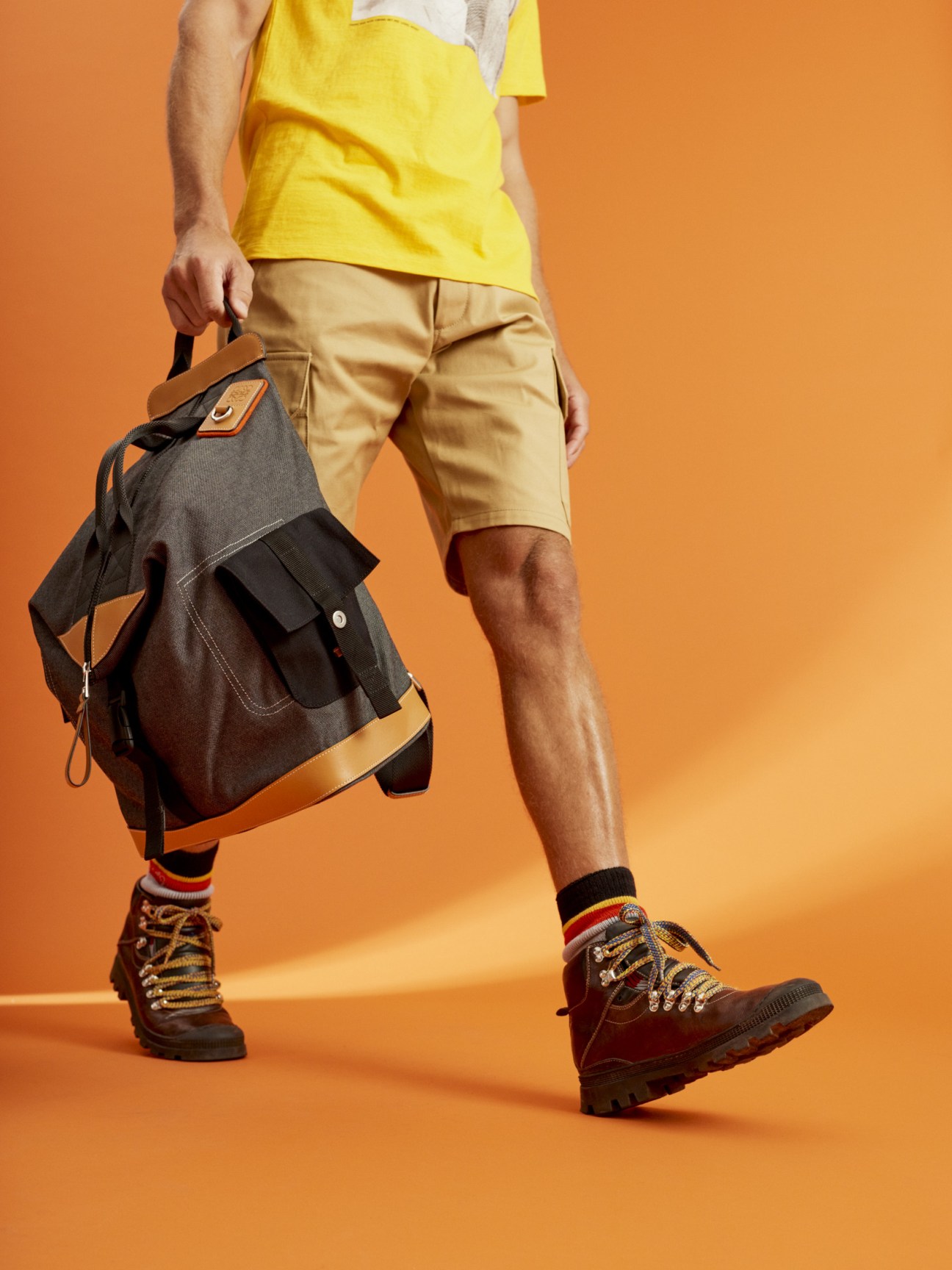 Man wearing yellow t-shirt, beige shorts, striped socks and hiking boots holding a canvas backpack with leather trim against an orange background.