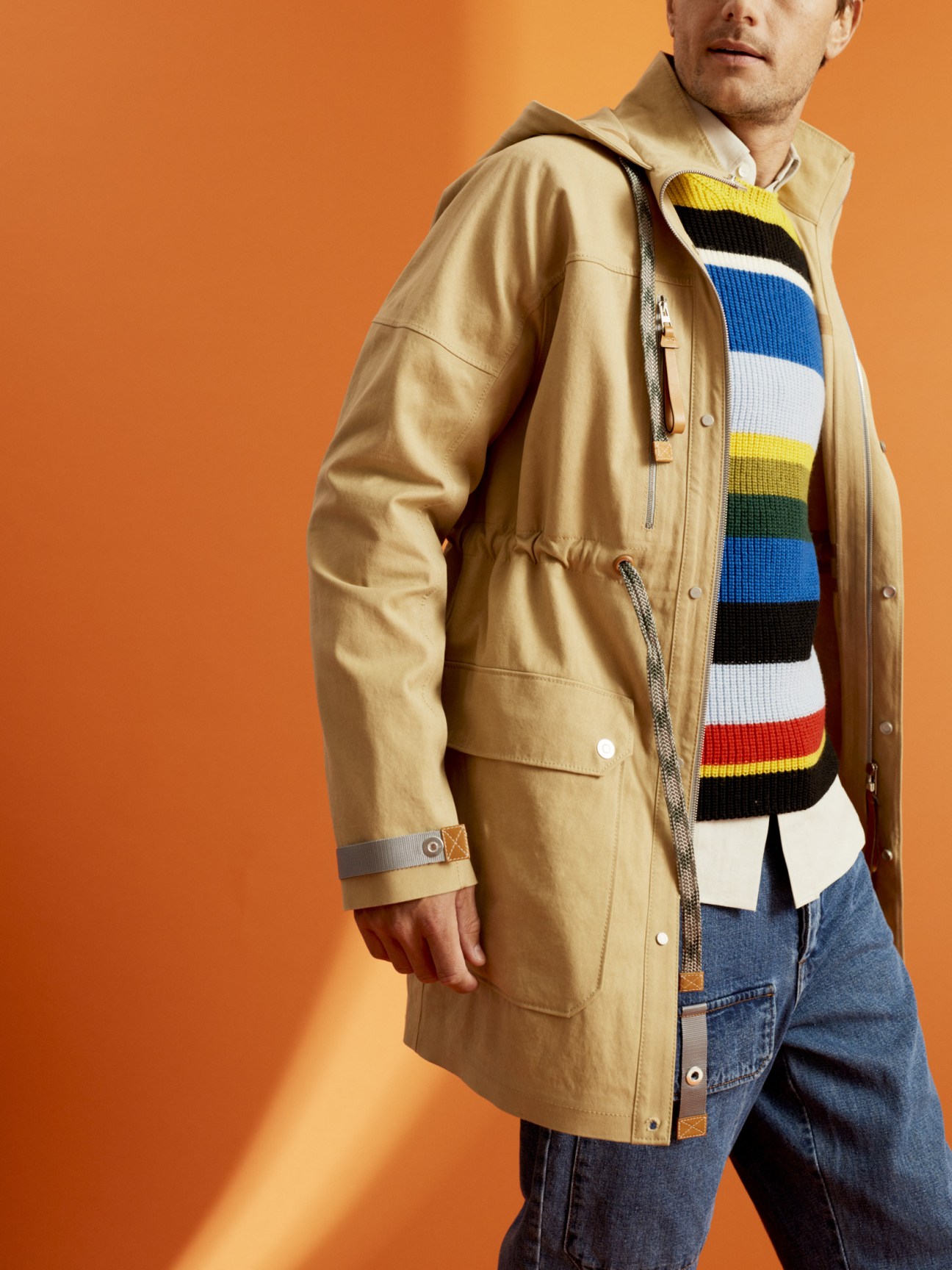 Man wearing a beige long parka, striped jumper, Oxford shirt and cargo jeans from Eye Loewe Nature against an orange background.
