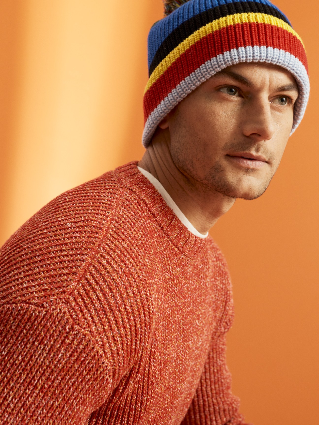 Man wearing Eye Loewe Nature striped beanie and red crewneck sweater against orange background.