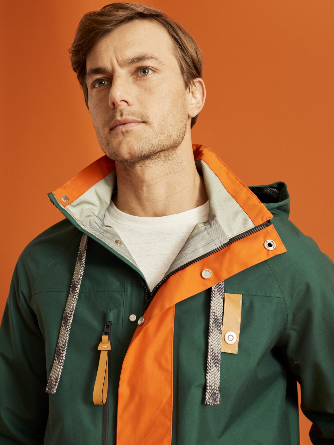 Man wearing green and orange parka over white t-shirt against orange background.