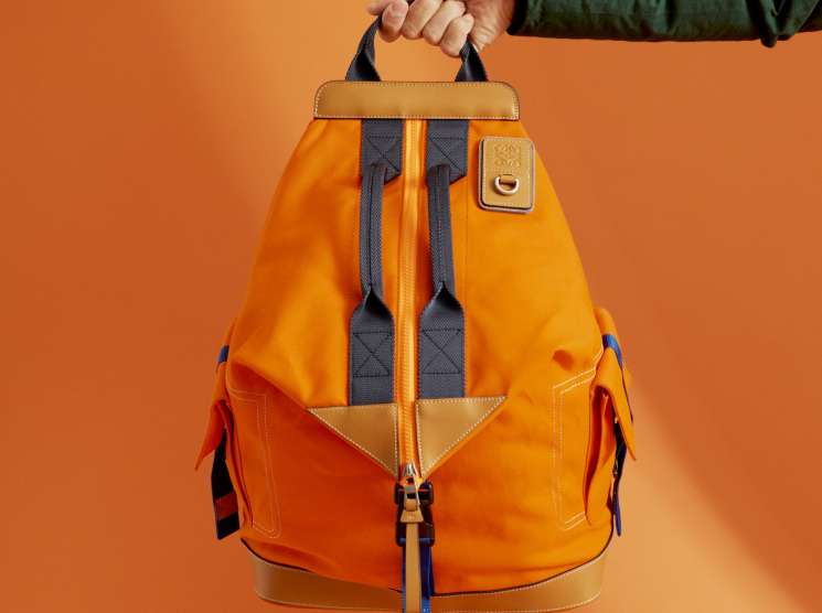 Person holding an Eye Loewe Nature orange canvas backpack with tan leather trim and navy straps against orange background.
