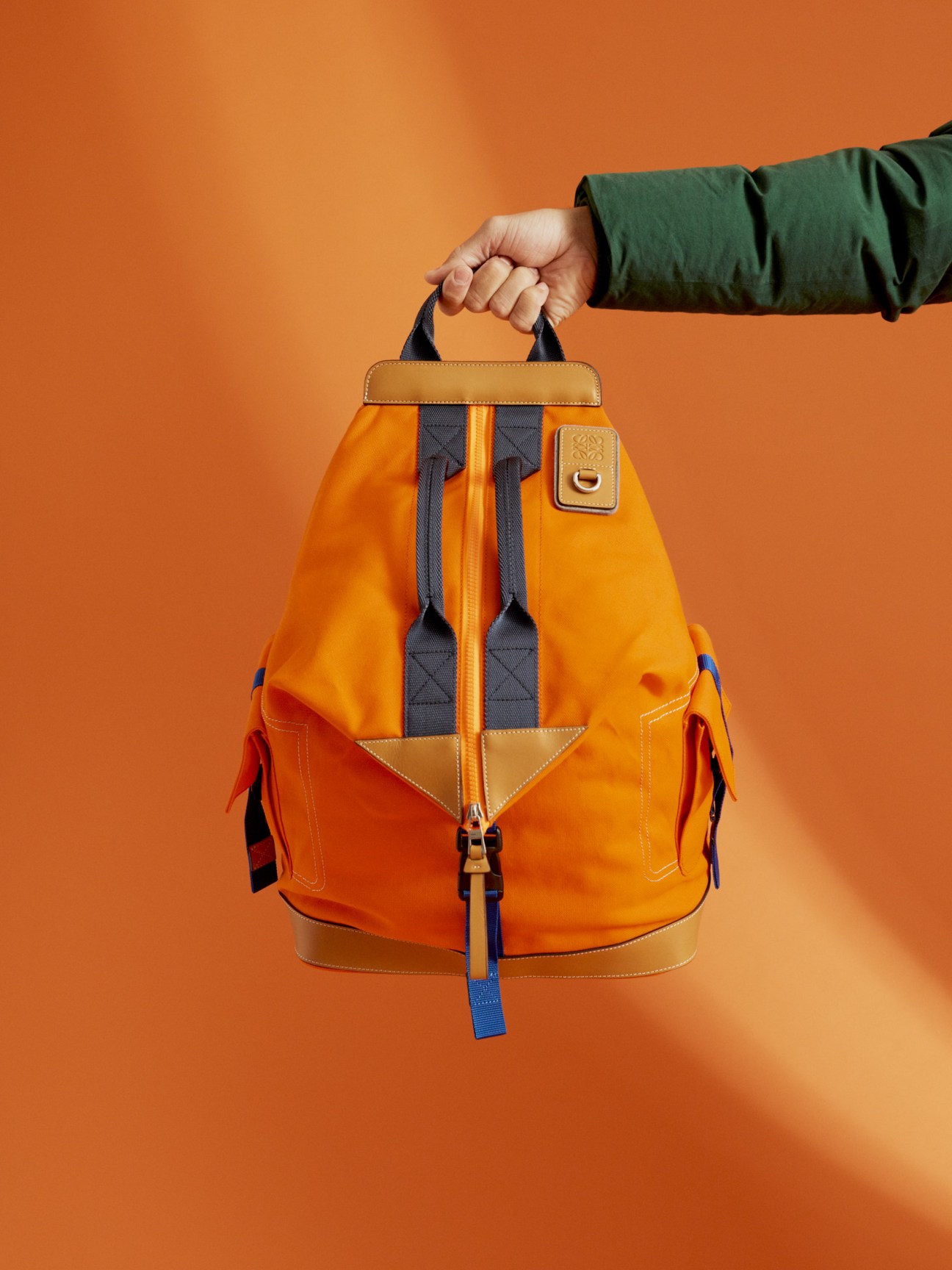 Person holding an Eye Loewe Nature orange canvas backpack with tan leather trim and navy straps against orange background.