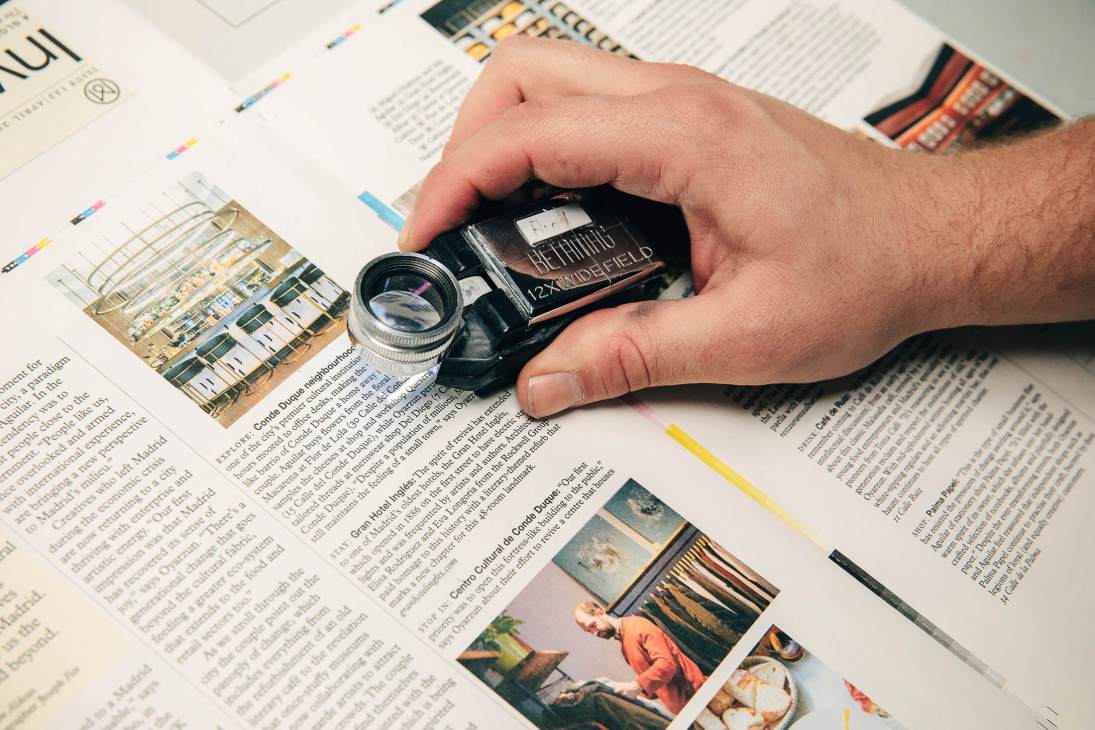 Hand holding a magnifying loop over magazine pages to inspect colour quality during printing production.