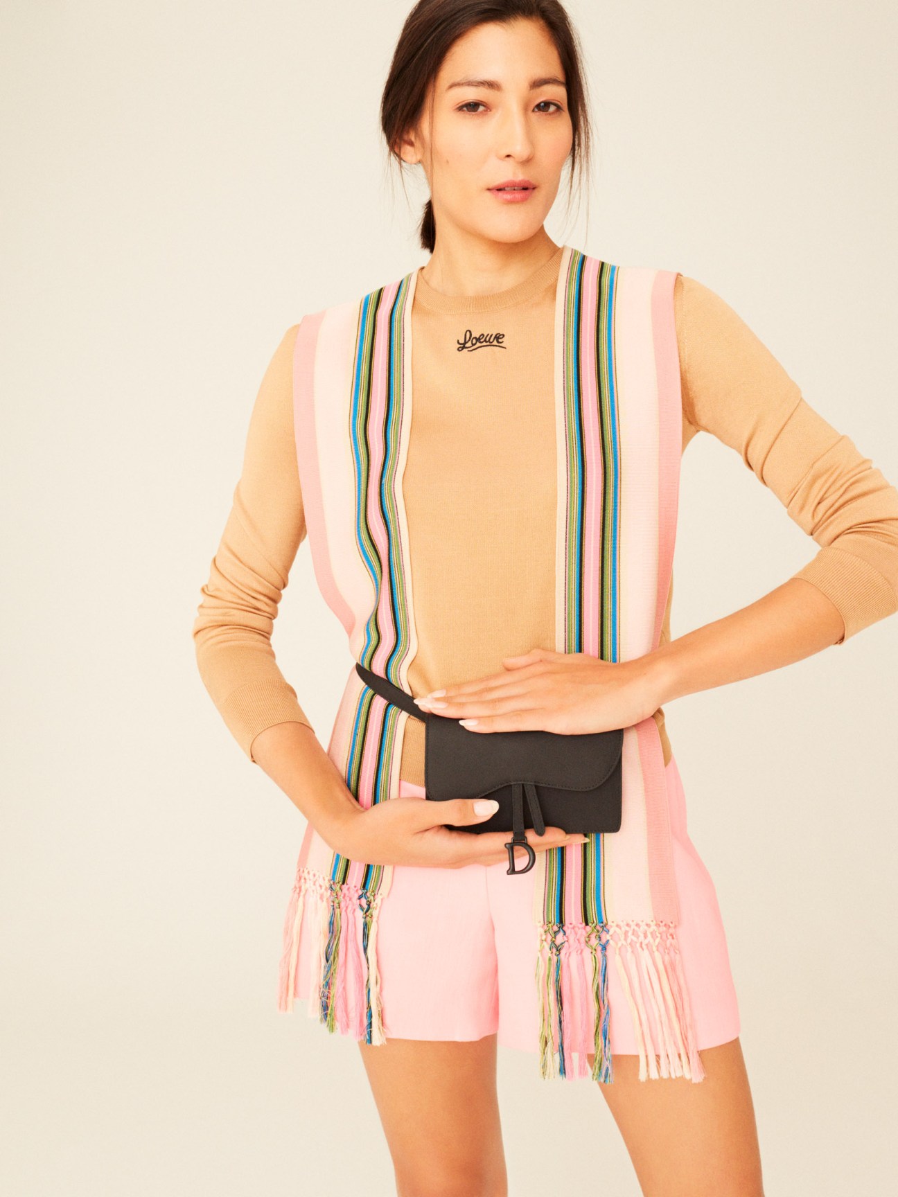 Woman wearing a peach jumper by Loewe with multicolored striped scarf and pink shorts, holding a black waist pouch by Dior.
