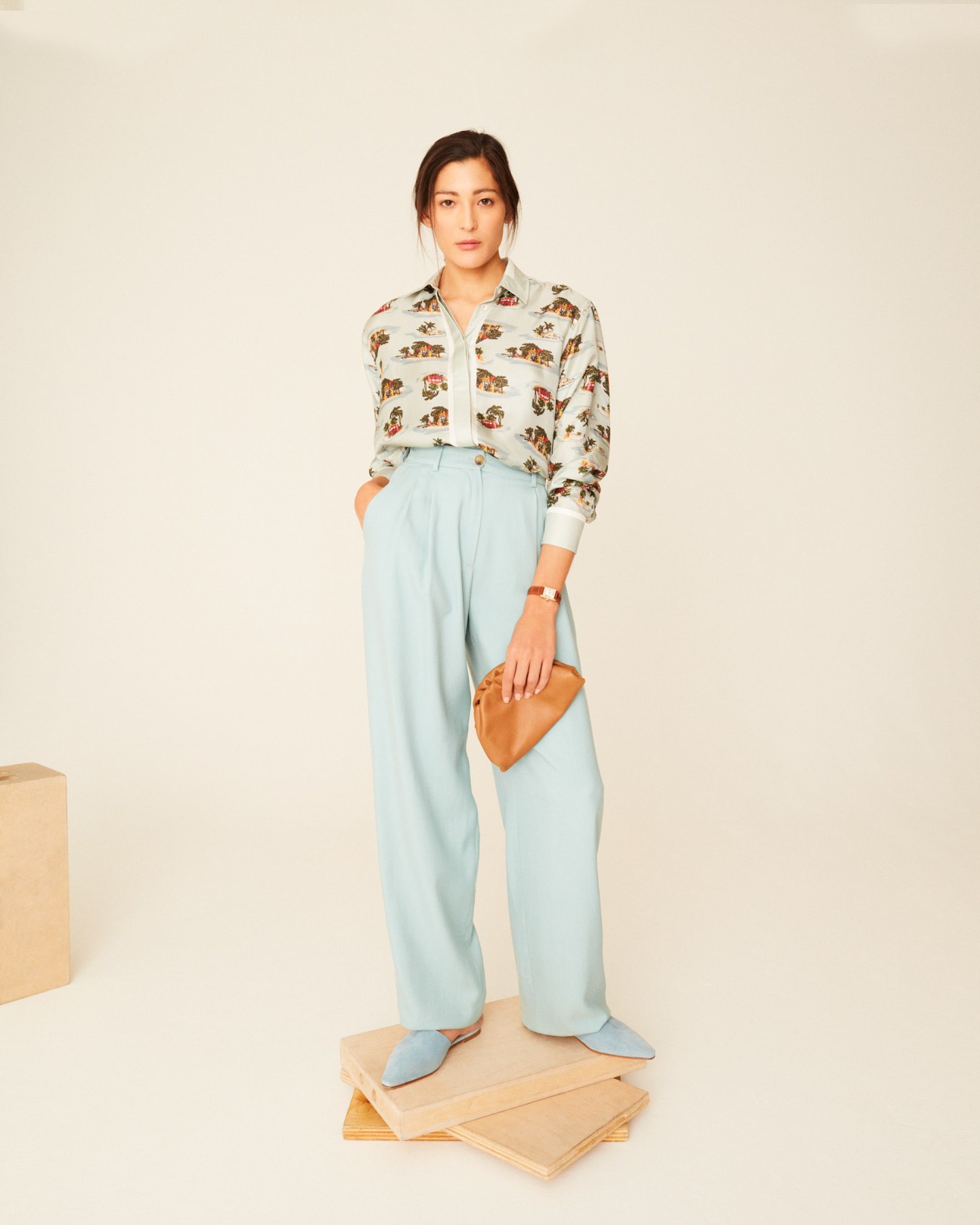 Woman wearing a Bottega Veneta patterned shirt, Bally light blue trousers, and holding a tan clutch.