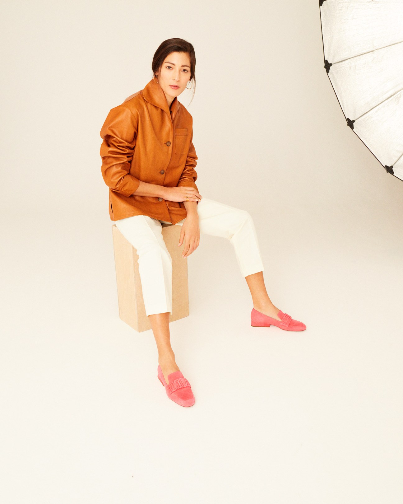 Woman wearing an orange Bottega Veneta shirt, white and beige striped trousers, and coral loafers sitting against a neutral background.