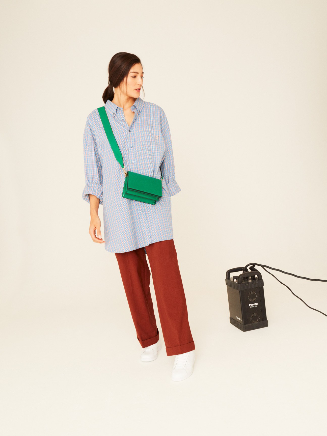 Woman in blue striped shirt and rust-colored trousers with green crossbody bag and white sneakers