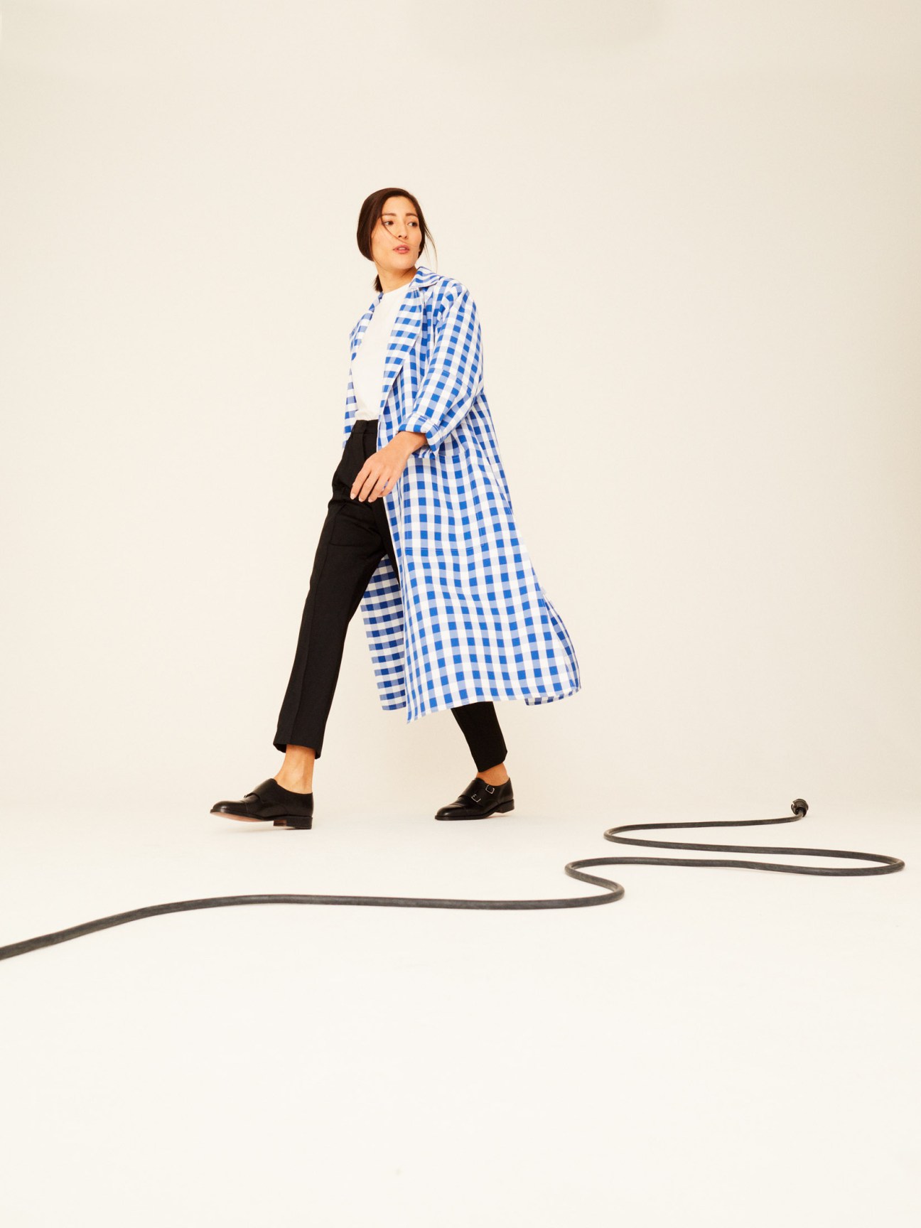 Woman wearing blue gingham coat, black trousers and shoes standing against cream background with dark cord.
