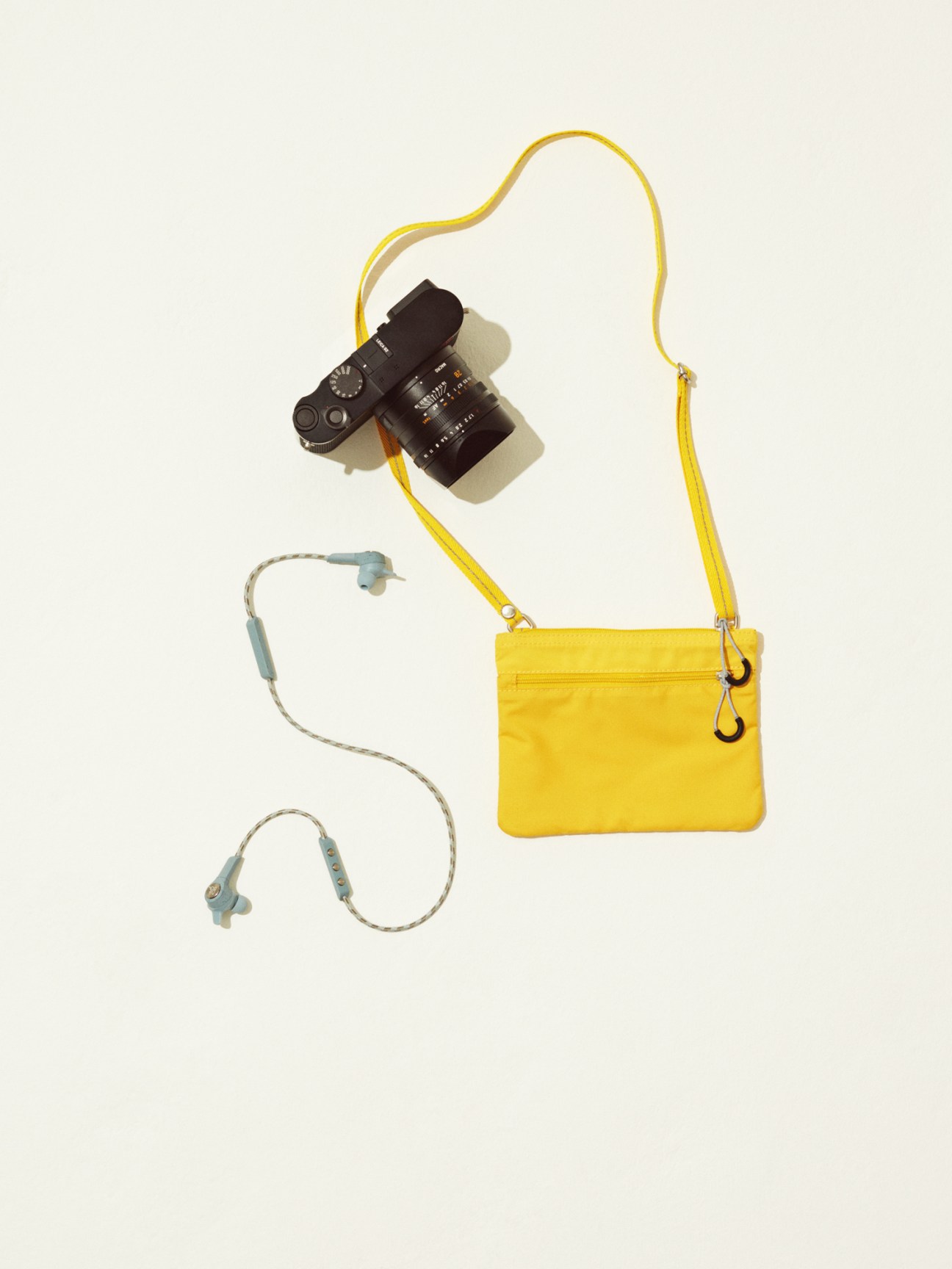 Yellow crossbody bag by Arket with Leica camera and Bang & Olufsen earphones arranged on neutral background.