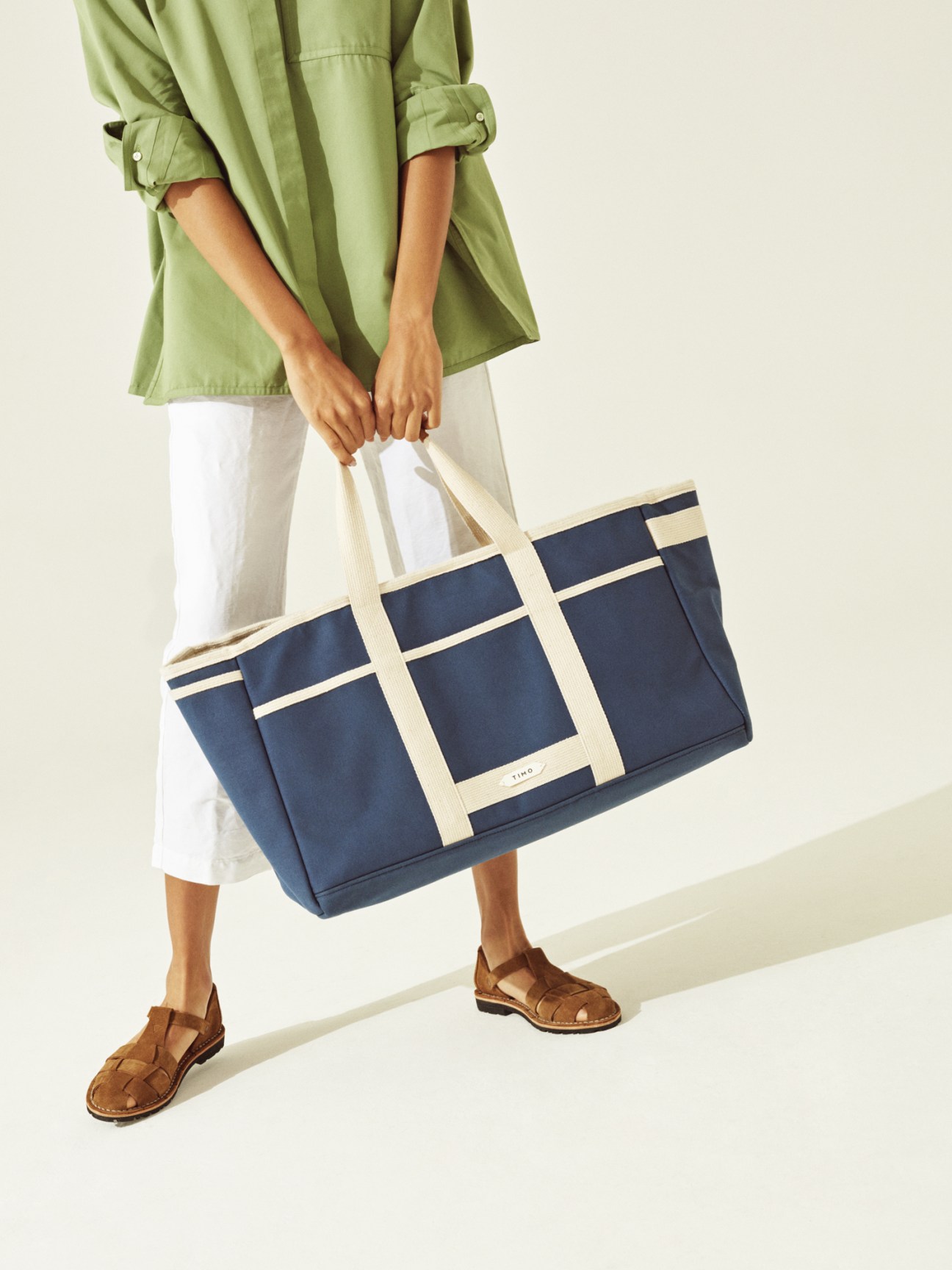 Woman wearing green shirt and white trousers holding navy and cream Timo tote bag with brown sandals.