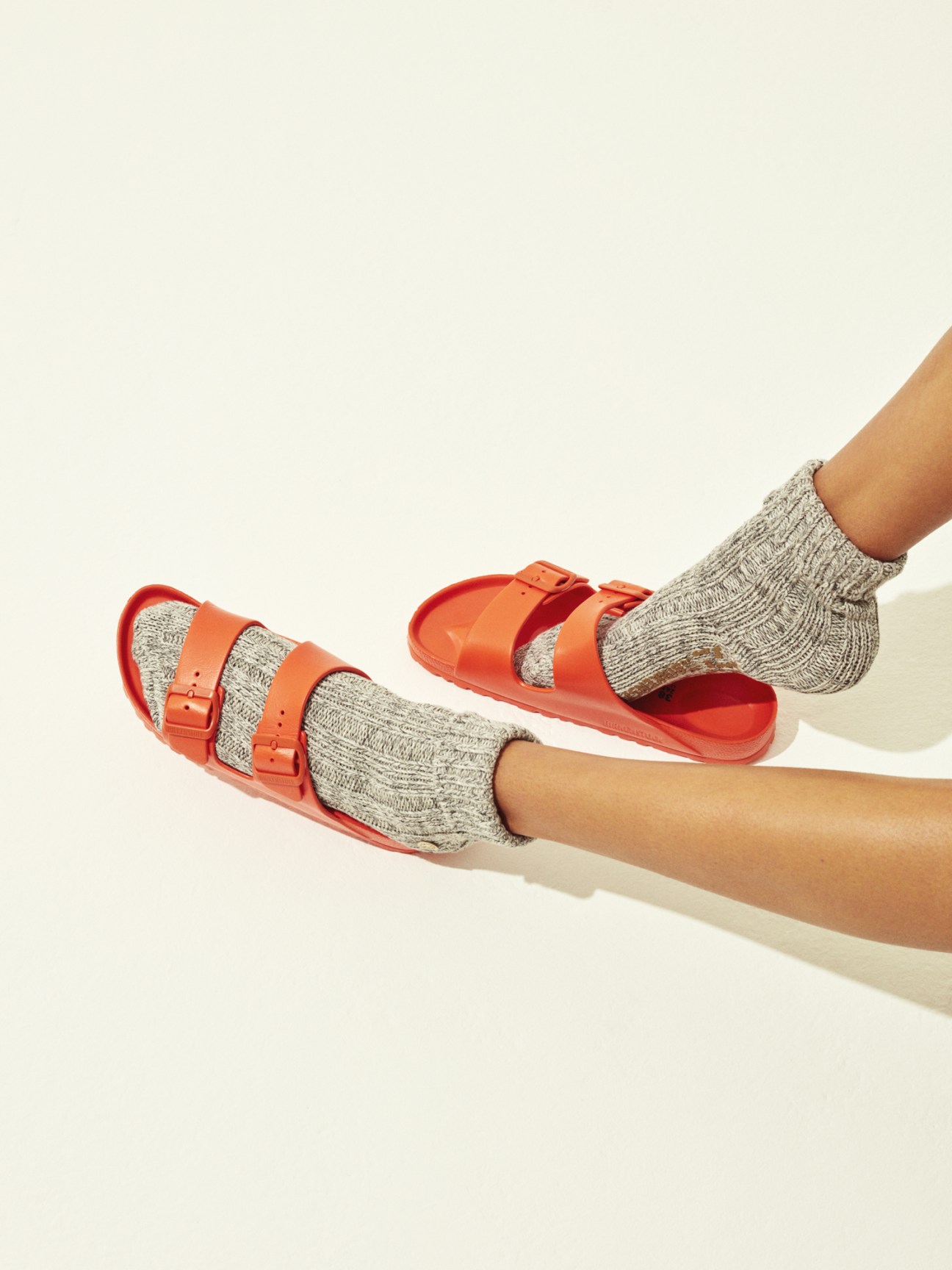 Red Birkenstock sandals worn with grey knit socks on two legs against a cream background.