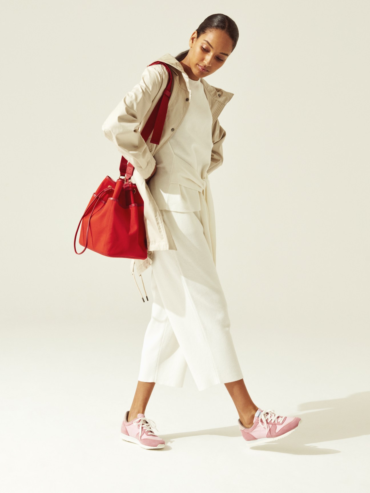 Woman wearing cream jacket and white trousers with pink sneakers, holding a red bucket bag.