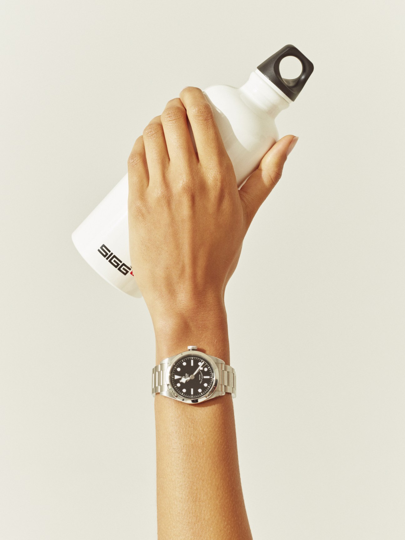 Hand holding a white Sigg water bottle and wearing a Tudor watch against a neutral background.