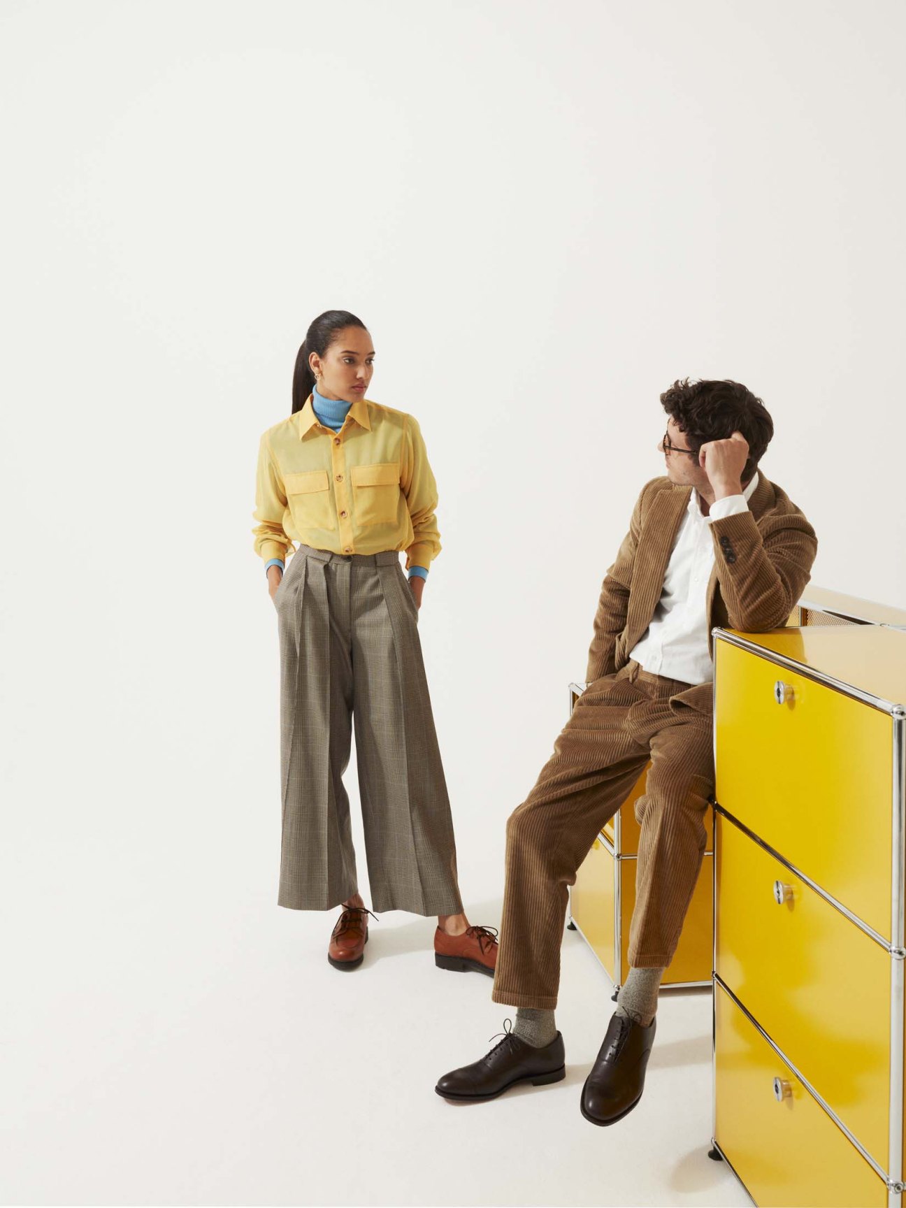 Man in brown corduroy suit and white shirt sits on yellow cabinet, wearing glasses.