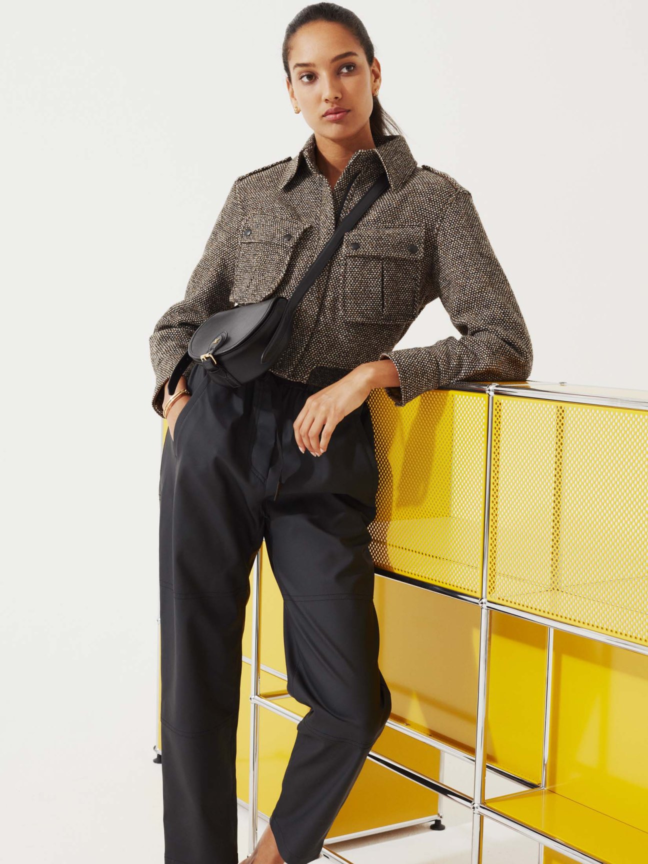 Woman in navy blazer and matching trousers with brown leather handbag leaning against yellow metal furniture.
