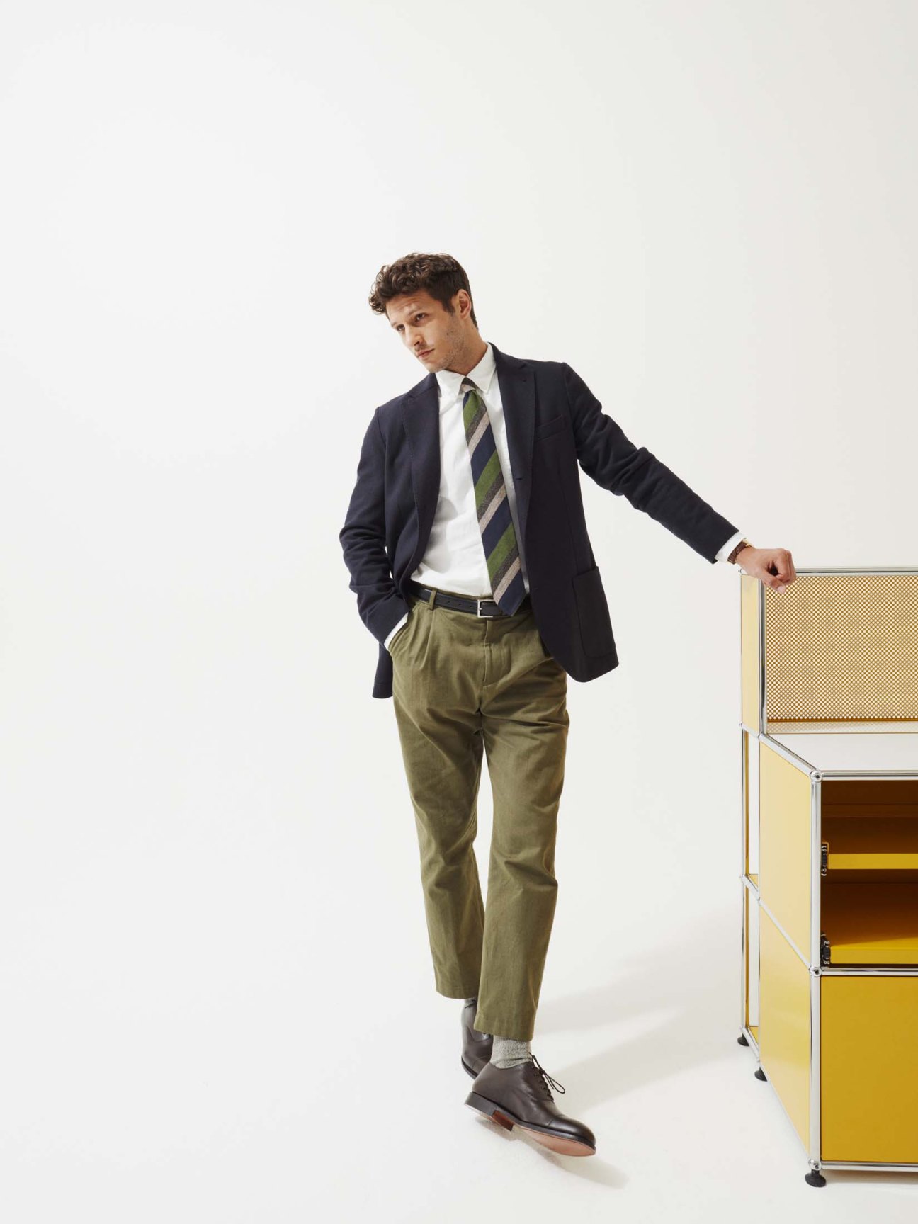 Man in navy blazer with white shirt and blue striped tie, leaning against yellow and beige shelving unit.