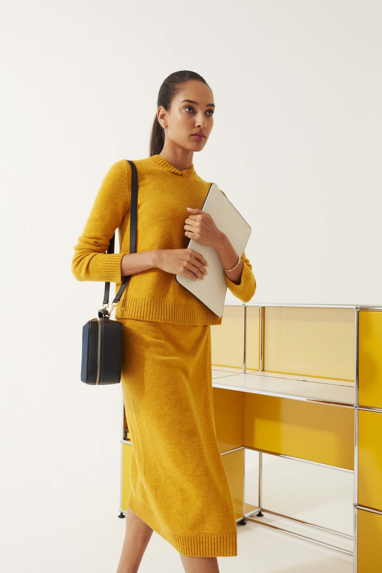 Woman in mustard-yellow Loro Piana jumper and midi skirt holding cream leather pouch with black shoulder bag.