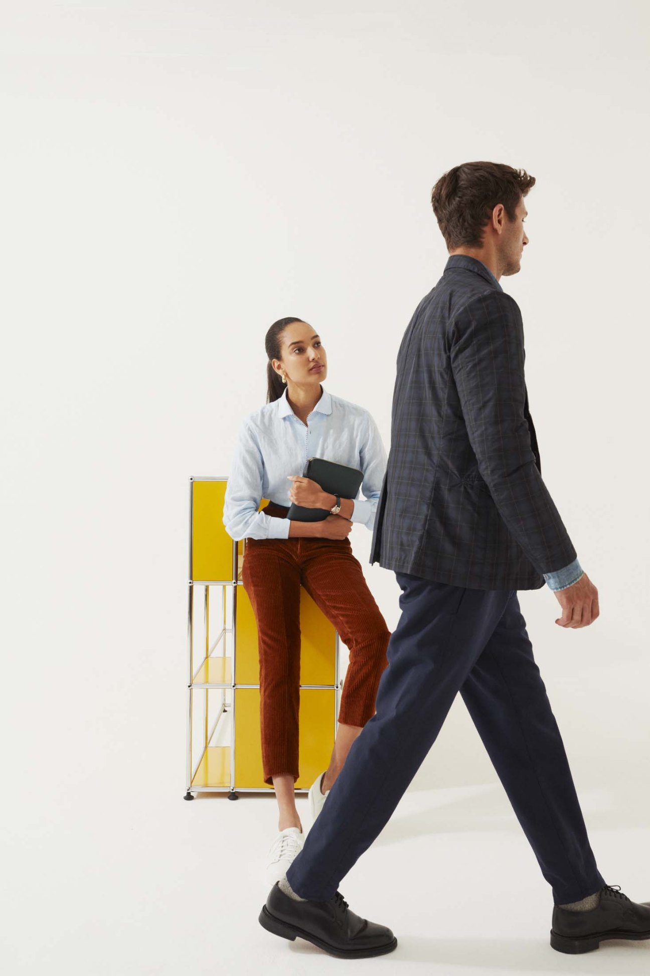 Woman in white shirt and rust trousers with black accessories beside man in charcoal blazer and navy trousers.
