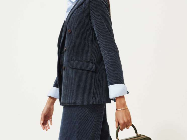 Woman in navy pinstriped blazer and trousers with white shirt holding olive green handbag.