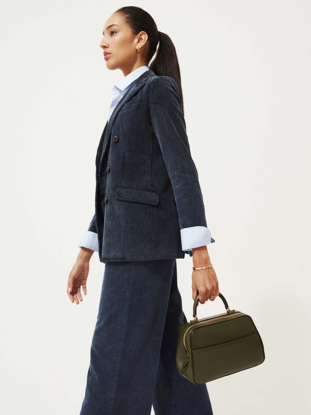 Woman in navy pinstriped blazer and trousers with white shirt holding olive green handbag.
