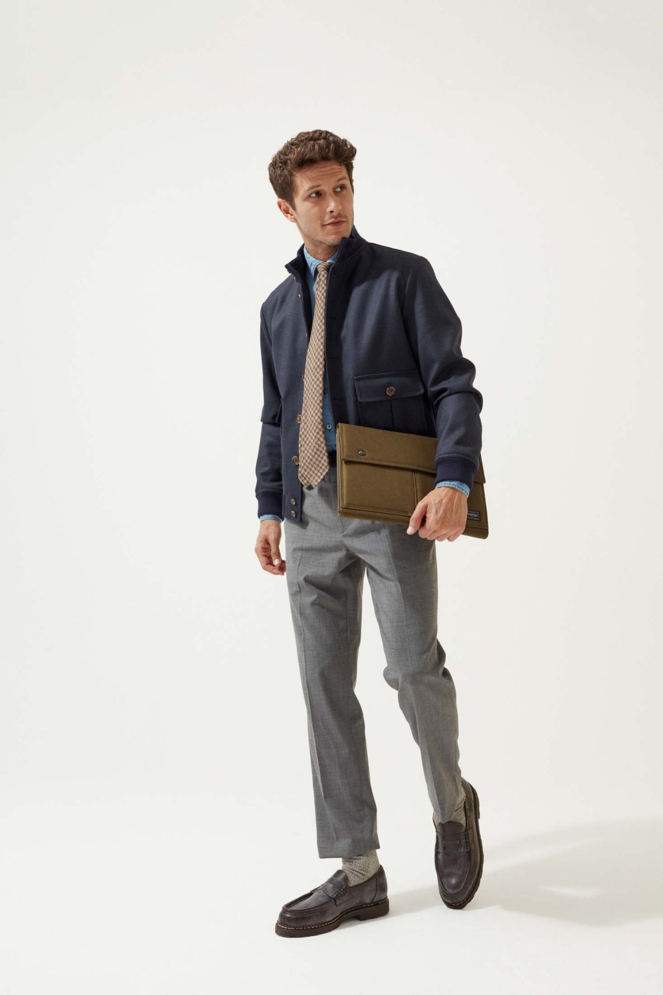 Man in navy blazer with patterned tie holding brown leather laptop case over grey trousers.