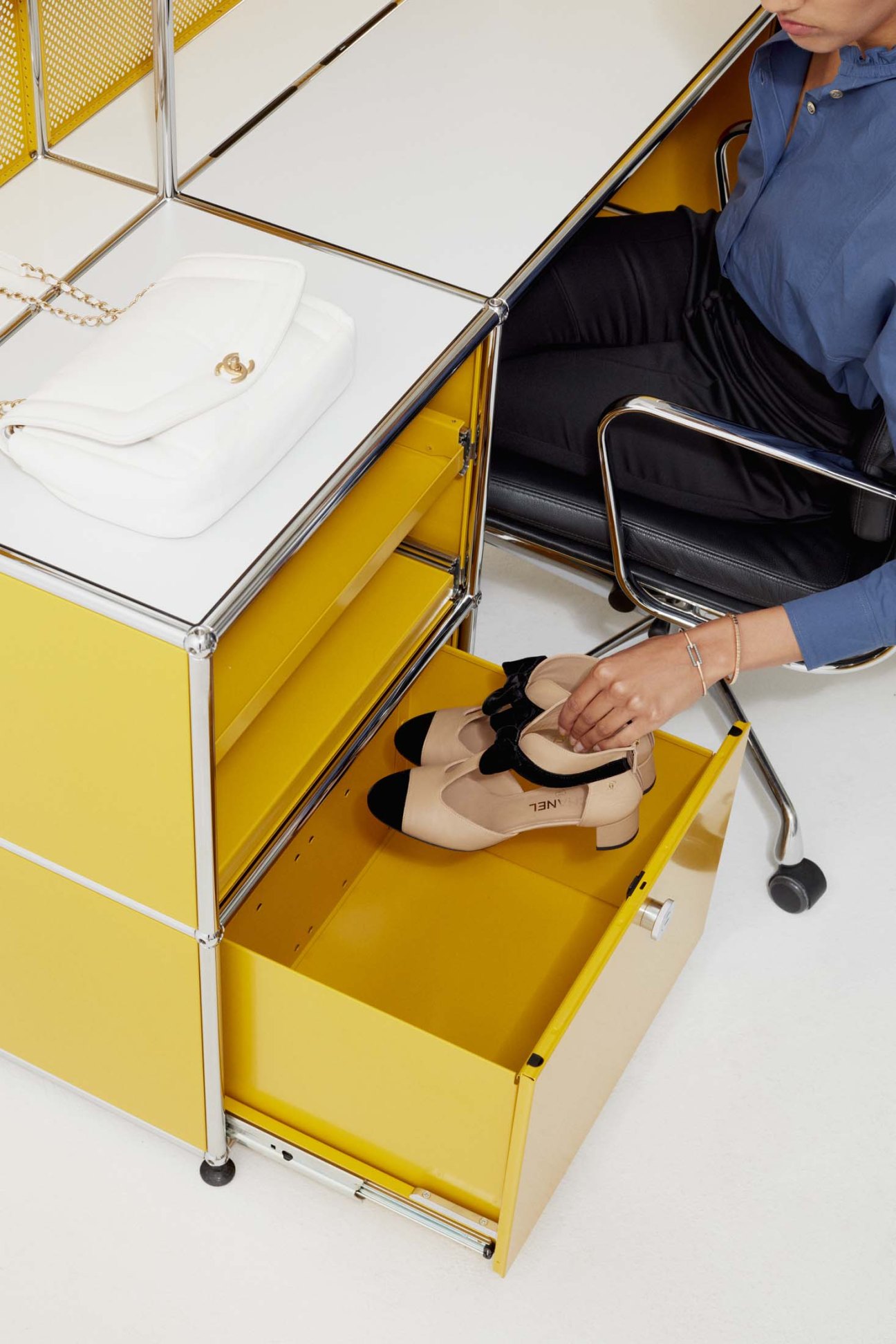 Woman in white shirt and navy trousers opening yellow storage unit drawer displaying Chanel bag and shoes