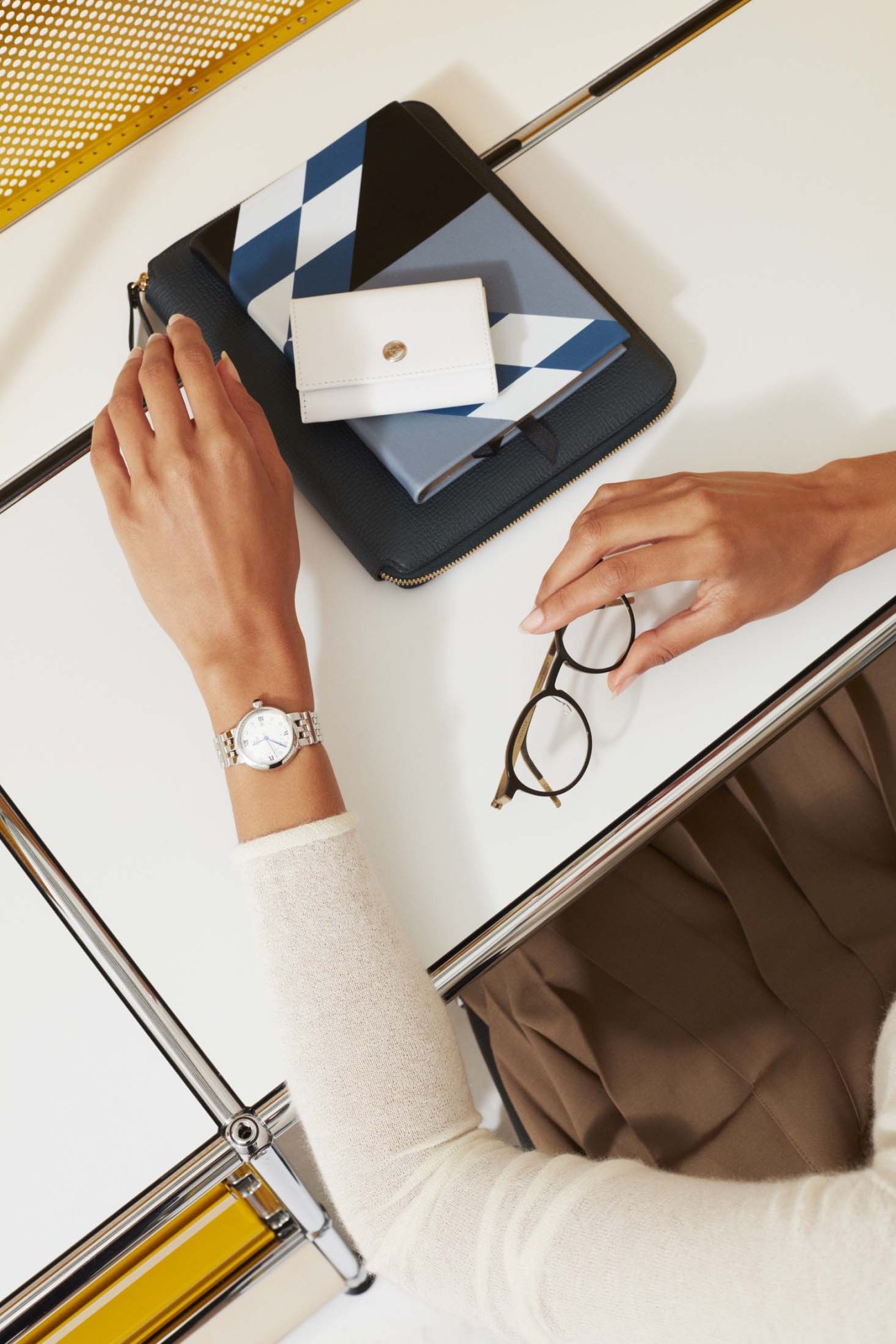 Woman's hands holding Smythson diaries, Ettinger coin purse, Tudor watch and Lindberg glasses on black wallet.