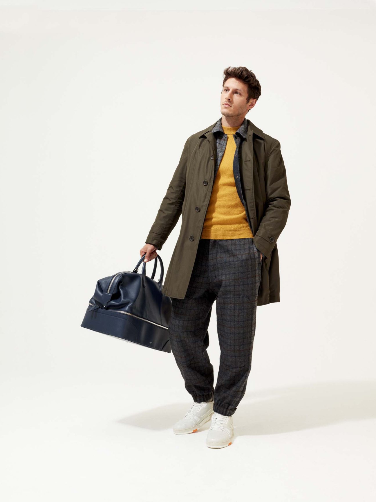 Man in navy coat and cream jumper holding navy leather bag wearing white trainers.