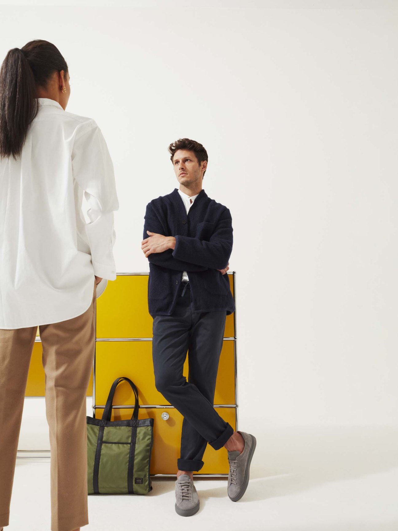 Woman in white shirt and tan trousers facing man in navy blazer leaning against yellow USM furniture