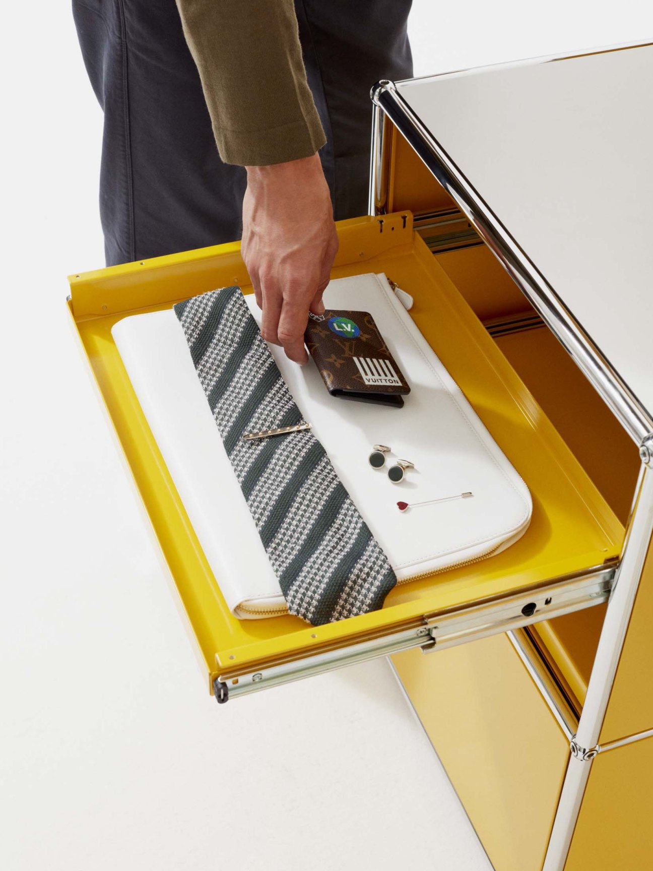 Man's hand placing a Louis Vuitton card case into a yellow drawer containing a Closed jumper, Aspesi trousers, Bigi tie, and Hermès accessories.