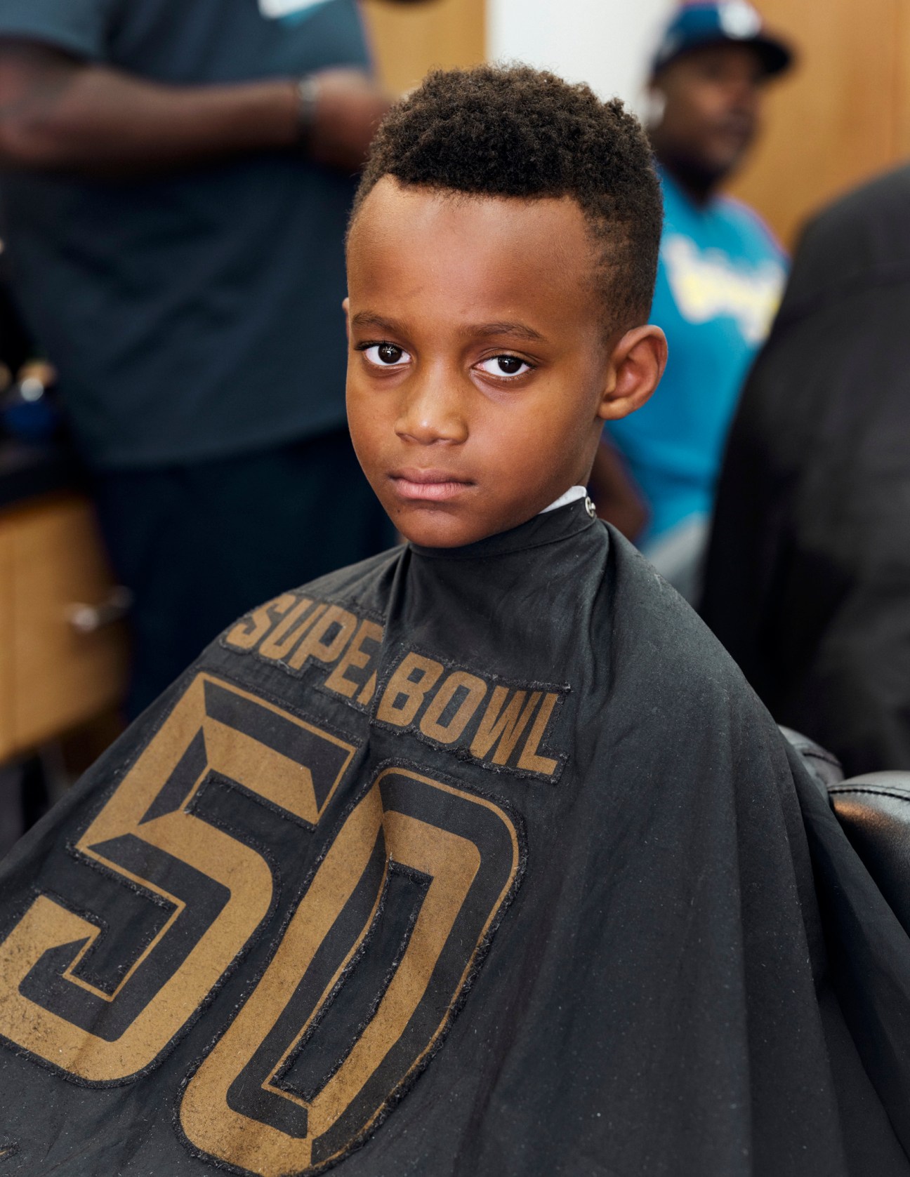 Young boy with sharp fade haircut wearing a black and gold Super Bowl 50 shirt.