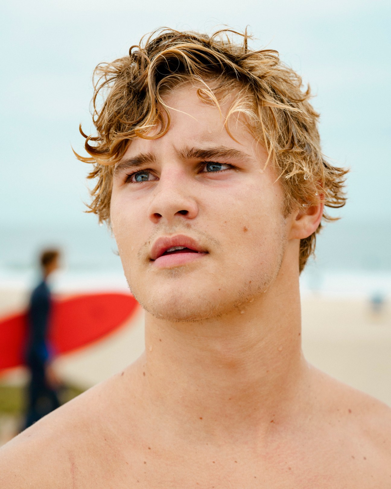 Young man with shoulder-length wavy blonde hair standing shirtless on a beach with surfers in background.
