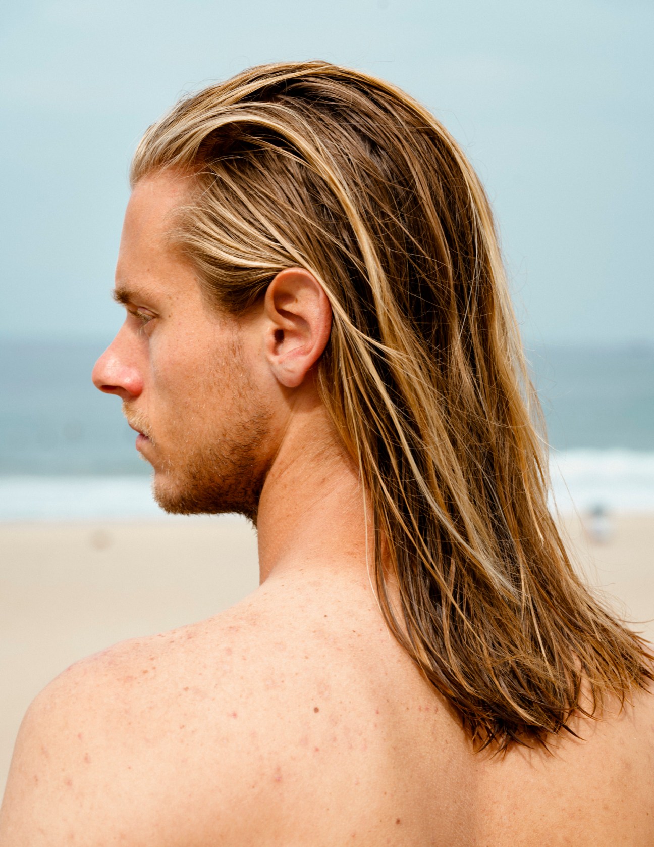 Clayton, a man with long shoulder-length blonde hair and facial hair, photographed in profile against a beach backdrop.