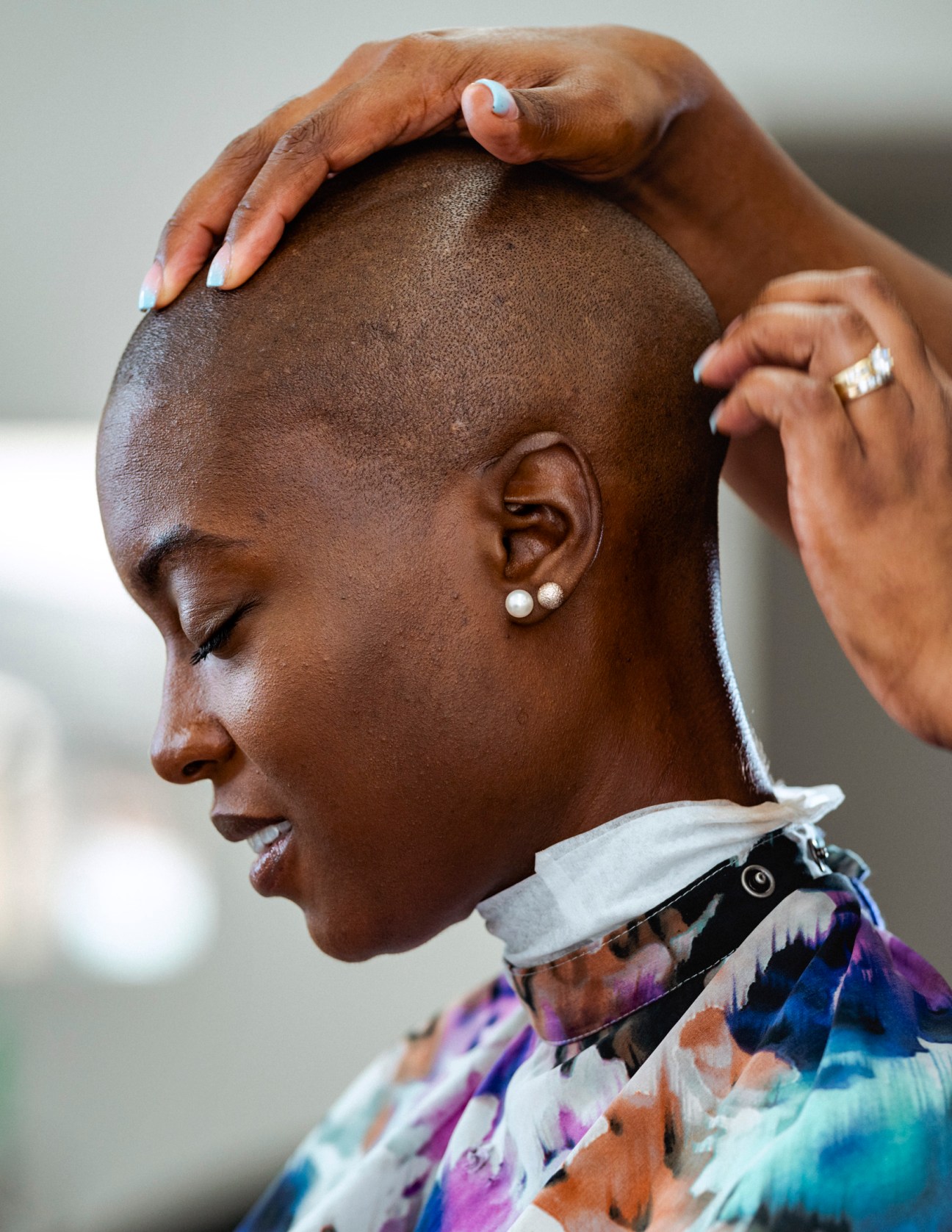 Woman with closely shaved head being touched by hands with white nail polish and gold rings.