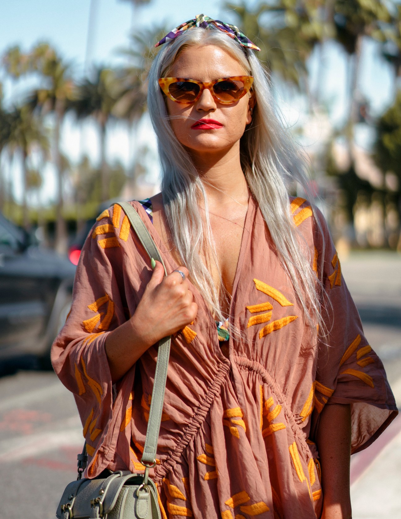 Woman with long silver hair wearing orange-tinted sunglasses and a patterned rust-colored shirt in sunlight with palm trees behind her.