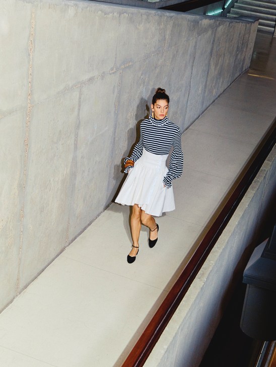 Model wearing striped rollneck jumper, white skirt and black shoes by Chanel in a concrete corridor.