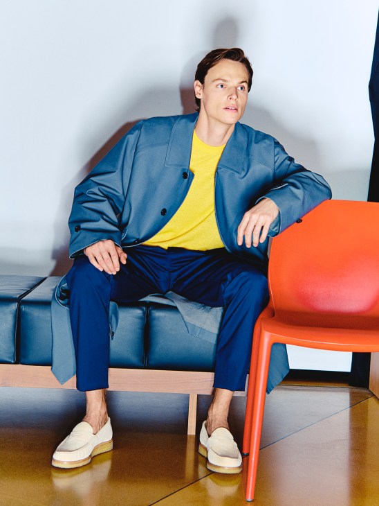 Man in blue blazer and navy trousers with yellow shirt and white shoes sits on modern furniture.