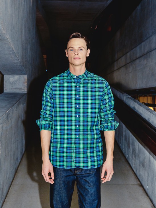 Man in green and blue plaid shirt by Scye Basics and Arket jeans standing in concrete corridor.