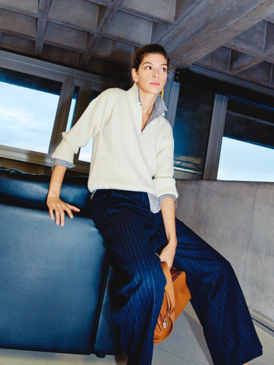 Model wearing cream CristaSeya jumper, Celine shirt and earrings, Patou trousers, and Hermès bag seated on blue seating.